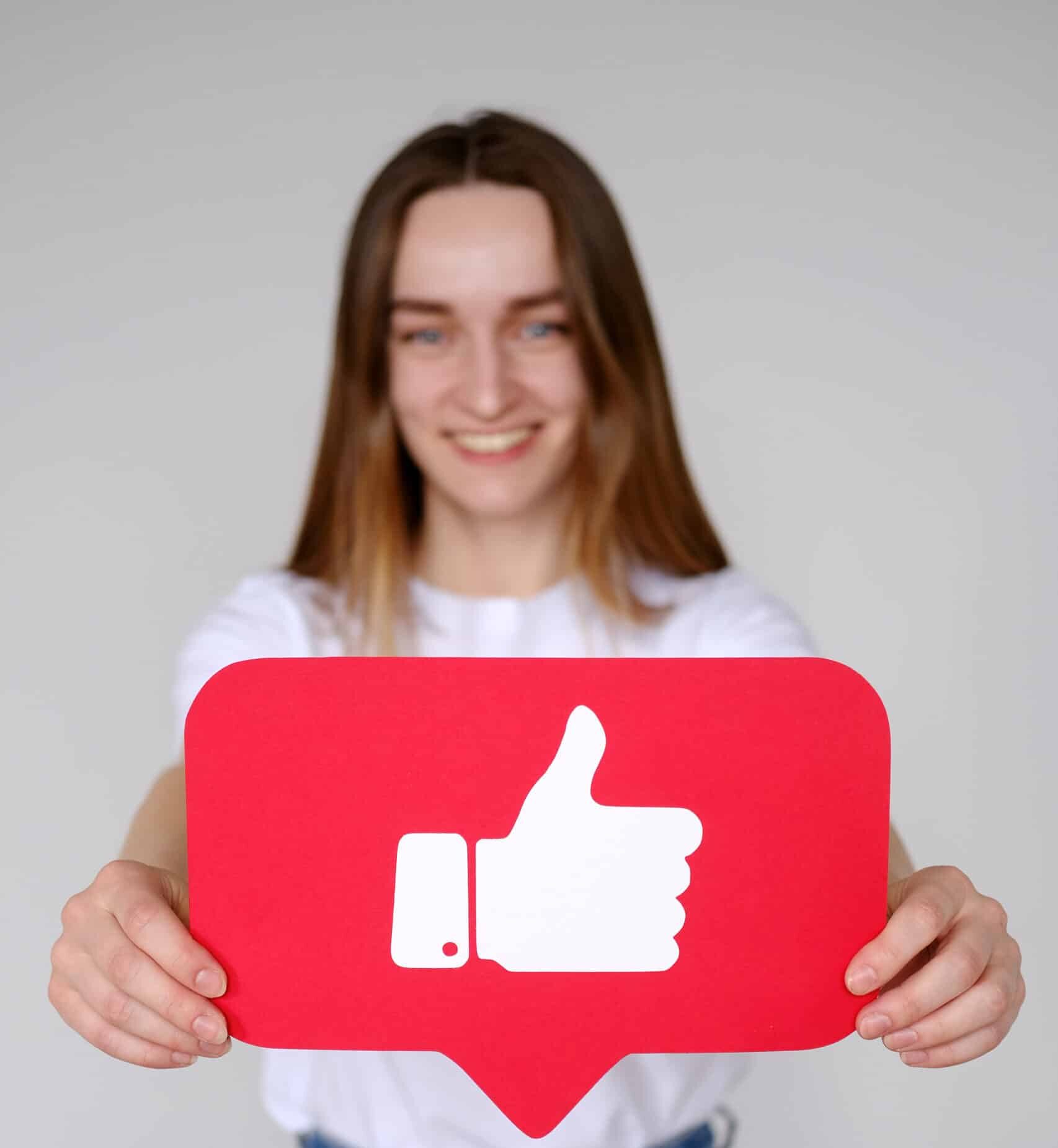 Cheerful carefree woman holding Like button icon of social media. Like button of social media. Smiling beautiful caucasian woman holding Like button icon, recommending to follow and share blog with interesting content, wearing white T-shirt.