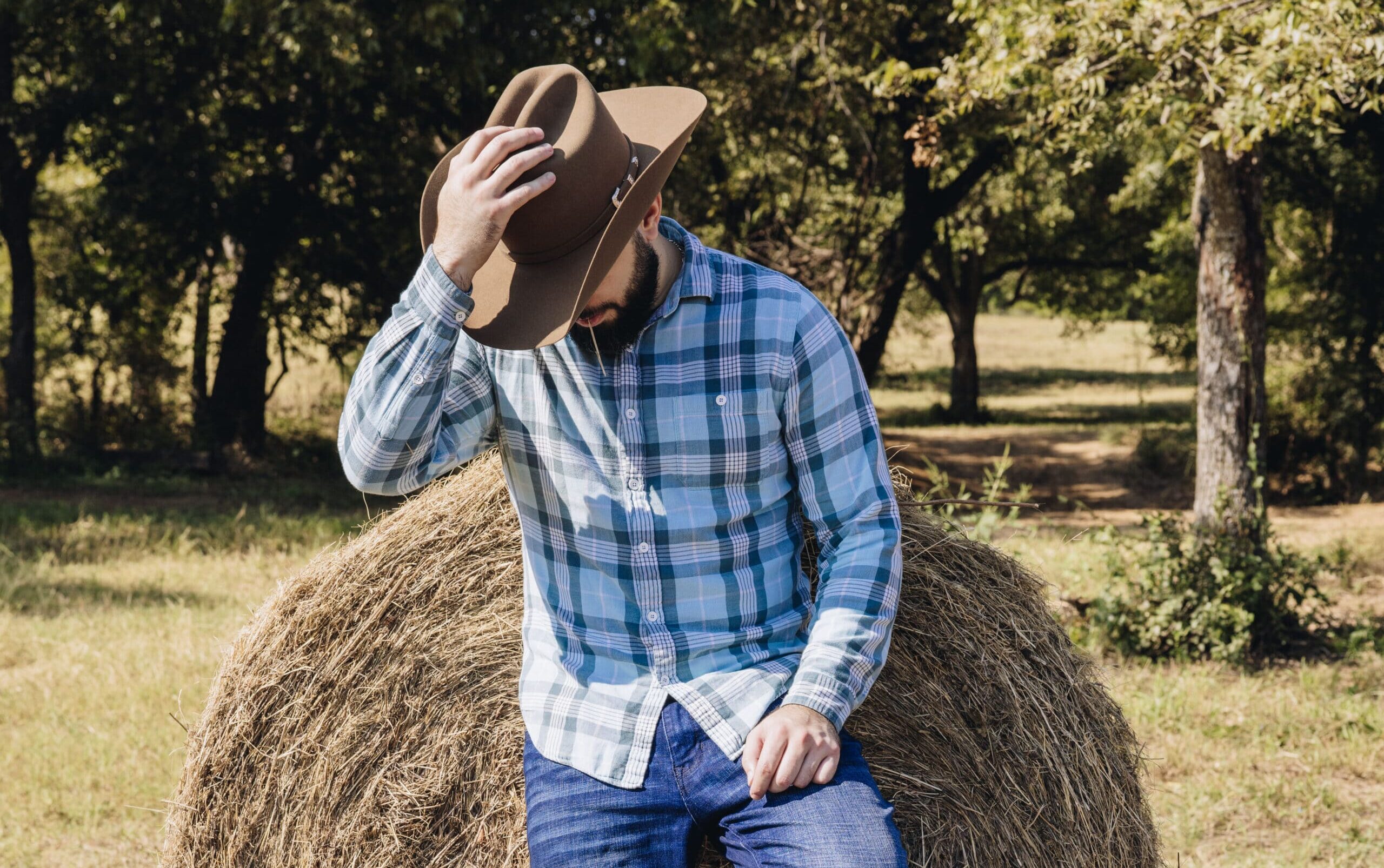 Man in plaid shirt and cowboy hat posing on hay bale in rustic setting
