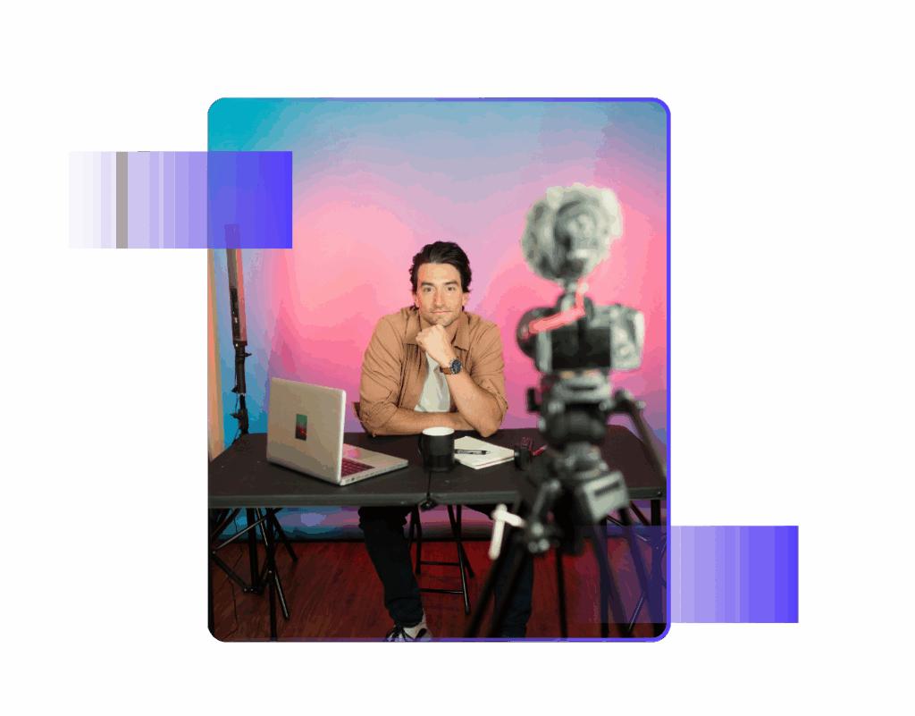 Influencer sitting at a desk with a laptop and camera setup recording a video against a pink and blue background.
