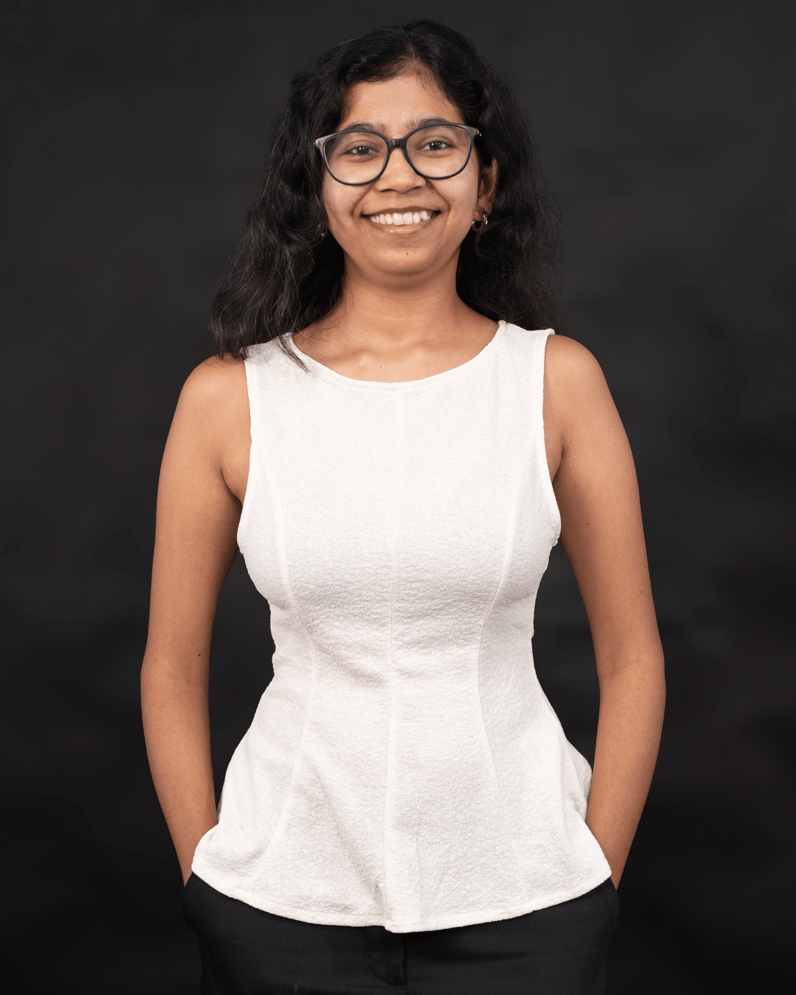 Young woman with glasses and a white sleeveless top smiling against a dark backdrop.
