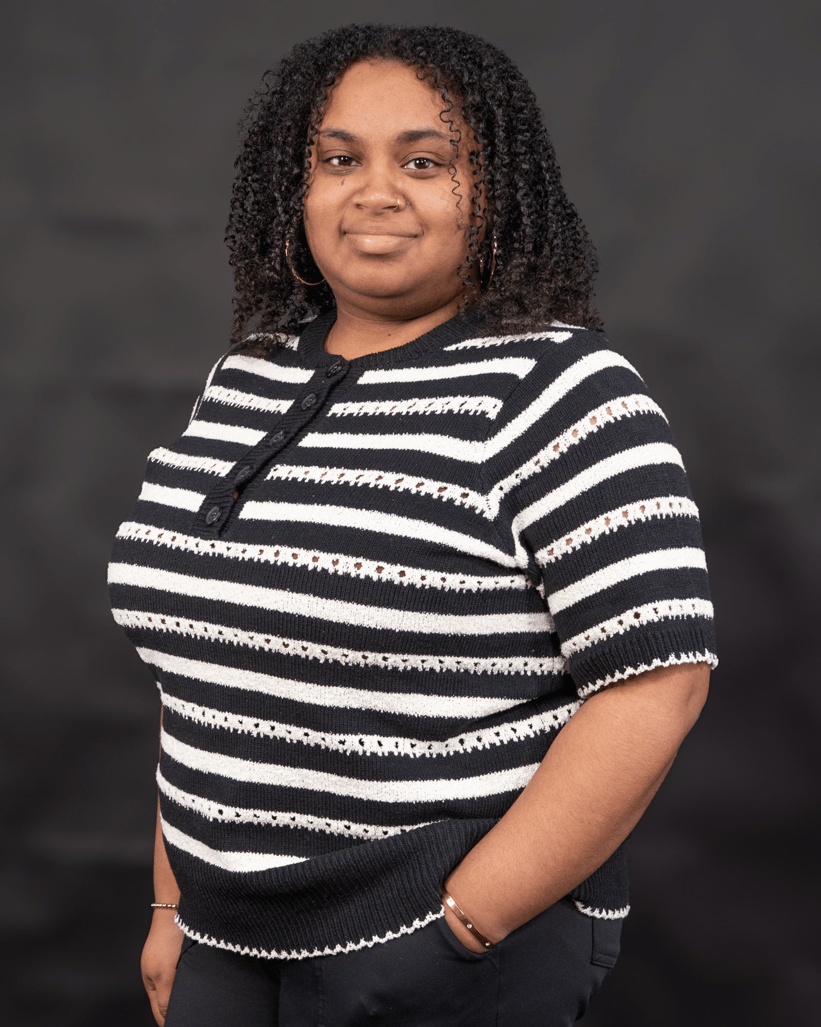Portrait of a woman with curly hair wearing a navy and white striped knit sweater, standing against a dark backdrop with a neutral smile.