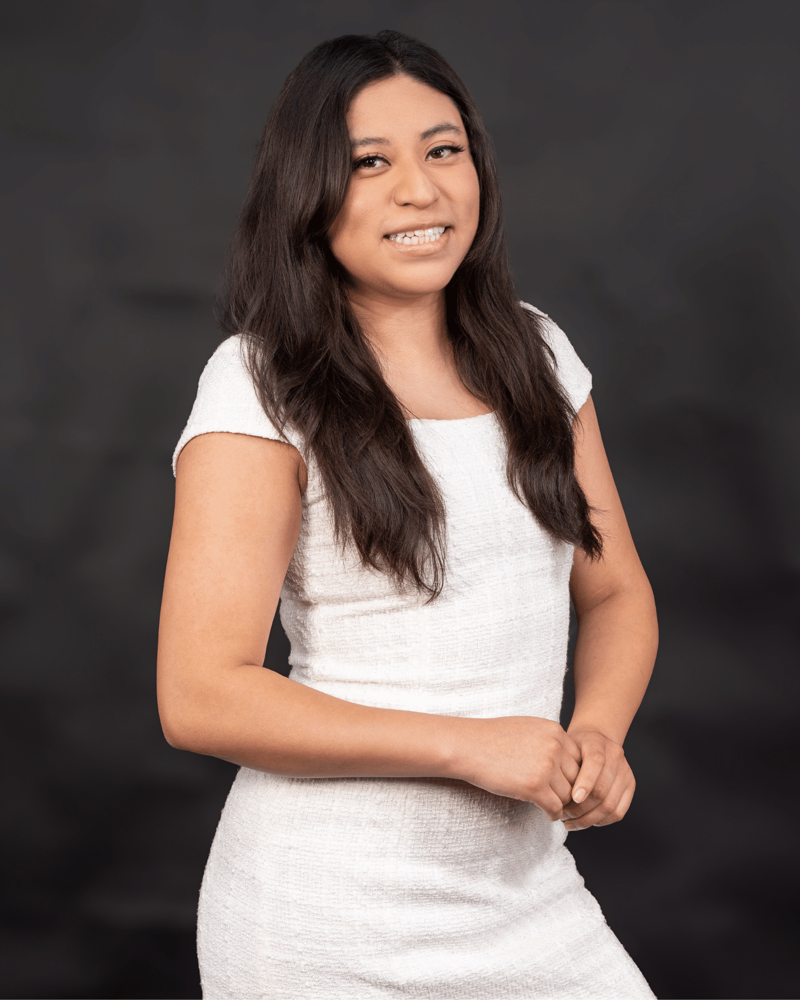 Portrait of a young woman with long dark hair, wearing a white textured dress, smiling softly against a dark studio backdrop.